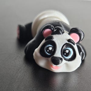 May include: A small, white and black panda figurine with pink ears and blue eyes. The panda is lying on its side with its head resting on its paws.