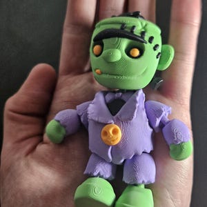 May include: A 3D-printed Frankenstein's monster figurine. The figure has a green head with black hair and stitches, orange eyes, and a purple jacket with an orange button. The hands and feet are green.