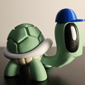 May include: A green turtle figurine with a white shell and a blue baseball cap. The turtle has a black eye and a green body.