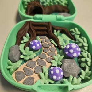 Puede incluir: Una escena de jard&iacute;n de hadas en miniatura dentro de un estuche verde. La escena incluye un toc&oacute;n de &aacute;rbol marr&oacute;n, un camino de piedra, rocas grises, follaje verde y setas moradas con lunares blancos. La mitad superior del estuche presenta un bosque borroso.