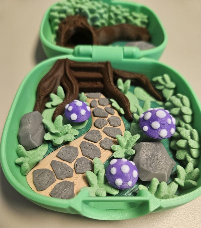 Puede incluir: Una escena de jard&iacute;n de hadas en miniatura dentro de un estuche verde. La escena incluye un toc&oacute;n de &aacute;rbol marr&oacute;n, un camino de piedra, rocas grises, follaje verde y setas moradas con lunares blancos. La mitad superior del estuche presenta un bosque borroso.