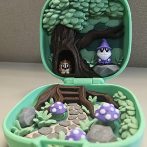 Puede incluir: Una escena de bosque en miniatura dentro de un estuche verde. La escena incluye un &aacute;rbol, setas, un camino y dos peque&ntilde;as figuras: un erizo y un mago. Las setas son moradas con puntos blancos. El estuche est&aacute; abierto, revelando el interior detallado.