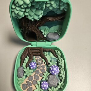Puede incluir: Un juego de miniaturas verde menta, abierto para revelar una escena de bosque. En el interior, un camino conduce a trav&eacute;s de un paisaje con &aacute;rboles, rocas y setas moradas con lunares. El juego se cierra con un bot&oacute;n morado.