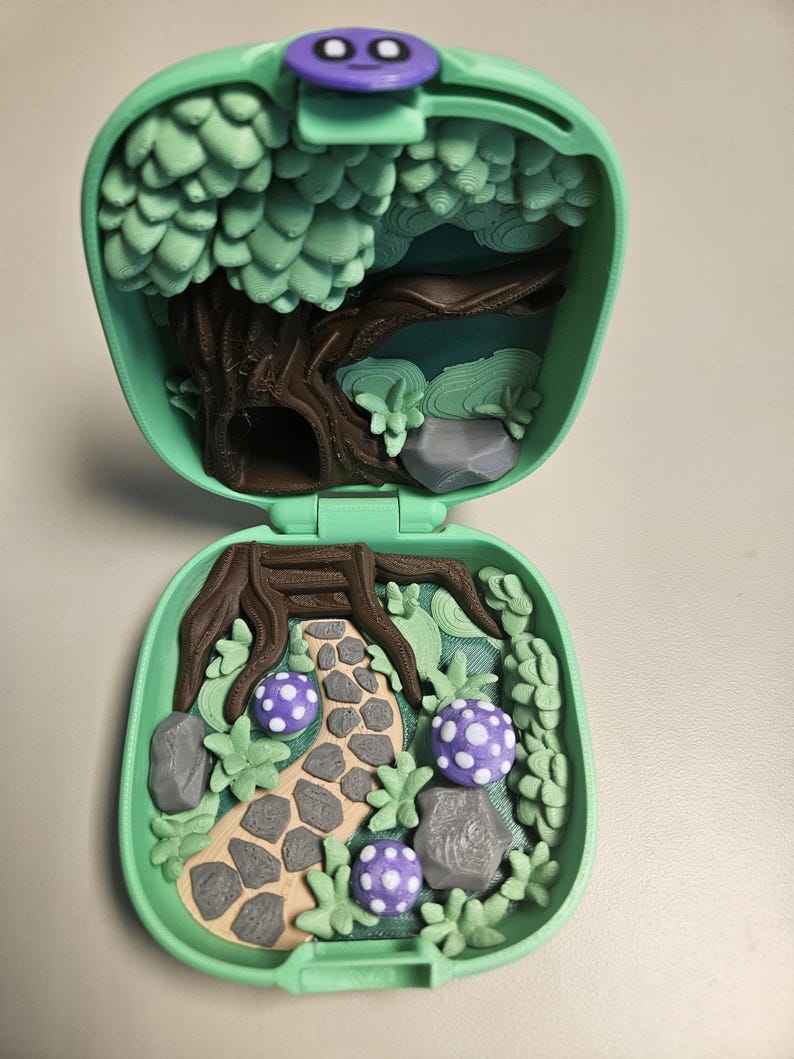 Puede incluir: Un juego de miniaturas verde menta, abierto para revelar una escena de bosque. En el interior, un camino conduce a trav&eacute;s de un paisaje con &aacute;rboles, rocas y setas moradas con lunares. El juego se cierra con un bot&oacute;n morado.