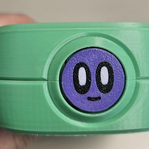 Puede incluir: Un recipiente verde y redondeado con un c&iacute;rculo morado en el centro. El c&iacute;rculo morado tiene una cara de dibujos animados con ojos blancos y una peque&ntilde;a sonrisa negra. El recipiente est&aacute; sostenido por una mano.