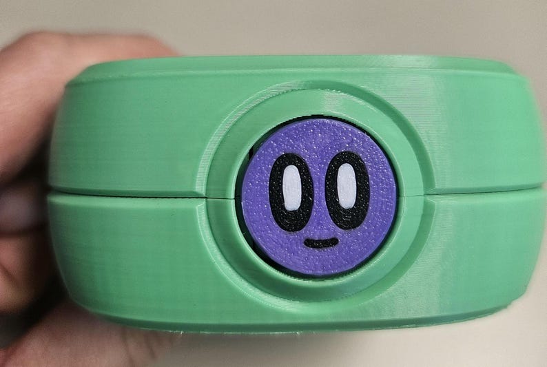 Puede incluir: Un recipiente verde y redondeado con un c&iacute;rculo morado en el centro. El c&iacute;rculo morado tiene una cara de dibujos animados con ojos blancos y una peque&ntilde;a sonrisa negra. El recipiente est&aacute; sostenido por una mano.