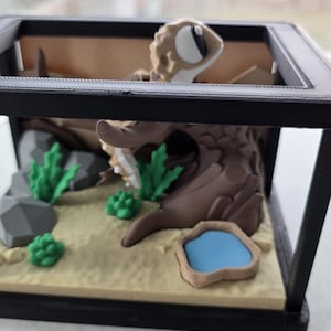 May include: A miniature terrarium scene featuring a gecko figurine. The scene includes a sandy base, a brown tree trunk, green plants, gray rocks, and a small blue pond. The terrarium is enclosed in a black frame.