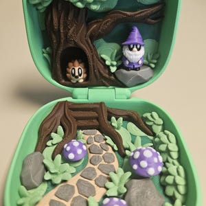 Puede incluir: Una escena de bosque en miniatura dentro de una concha verde. La escena incluye un &aacute;rbol, un gnomo, un erizo, setas y un camino de piedra. El gnomo lleva un sombrero morado y las setas tienen manchas blancas. La concha est&aacute; abierta, revelando el interior detallado.