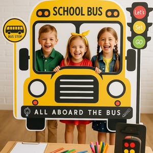 Può includere: Tre bambini posano per una foto davanti a un ritaglio di cartone di un autobus scolastico giallo. Il ritaglio ha le parole "School Bus" e "All Aboard The Bus" stampate su di esso. C'è un ritaglio di semaforo a destra del ritaglio dell'autobus. Il semaforo ha le parole "Let's", "Set" e "Go" stampate su di esso. Ci sono matite colorate e una cartellina con un foglio di carta bianco su un tavolo di legno davanti ai bambini.