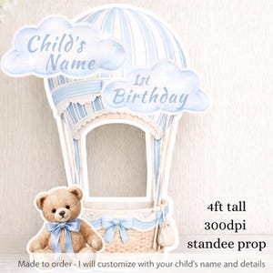 Pode incluir: Um suporte em forma de balão de ar quente azul e branco com o texto "Child's Name" e "1st Birthday". Um ursinho de pelúcia com uma fita azul está na frente. O suporte tem cerca de 122 cm de altura.