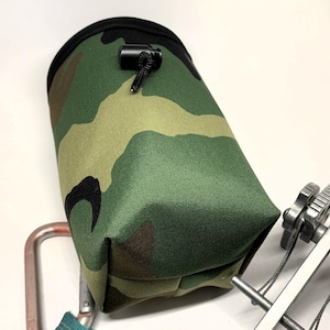 May include: A camouflage chalk bag for rock climbing, featuring a drawstring closure and a black rim. The bag is made of durable fabric and is designed to hold climbing chalk. A climbing carabiner is also visible.
