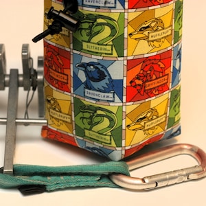 May include: A colorful fabric pouch with a drawstring closure and a carabiner clip. The pouch features a repeating pattern of the Hogwarts house crests: Gryffindor, Hufflepuff, Slytherin, and Ravenclaw.