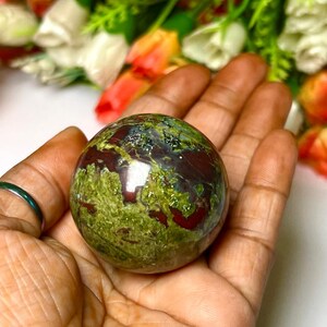 May include: A green and red jasper sphere, also known as a crystal ball, with a smooth, polished surface. The sphere is held in a hand.