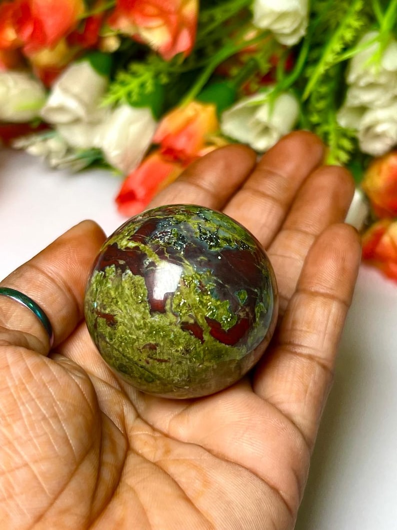 May include: A green and red jasper sphere, also known as a crystal ball, with a smooth, polished surface. The sphere is held in a hand.