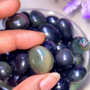 May include: A close-up of a hand holding a dark green and black iridescent gemstone. The gemstone is smooth and oval-shaped. The gemstone is surrounded by other similar gemstones in a bowl.