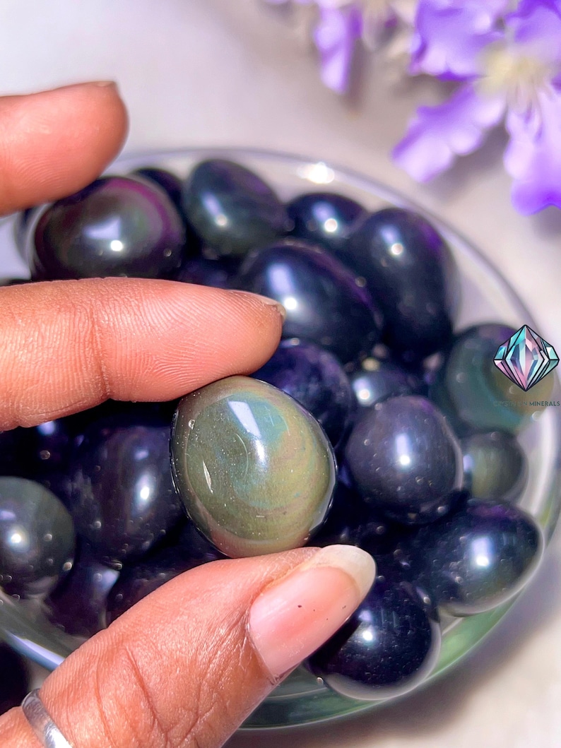 May include: A close-up of a hand holding a dark green and black iridescent gemstone. The gemstone is smooth and oval-shaped. The gemstone is surrounded by other similar gemstones in a bowl.