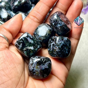 May include: A handful of polished, tumbled stones with a mix of dark blue, black, and white patterns. The stones have a glossy finish and are held in a hand. The image includes the text "CRYSTALS N MINERALS".