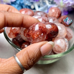 May include: A close-up of a hand holding a red and clear crystal tumbled stone. The stone has a red, white, and clear color with a sparkly, iridescent finish. The stone is surrounded by other tumbled stones in a glass bowl.