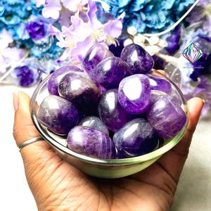 May include: A bowl filled with tumbled amethyst stones. The stones are a deep purple color and have a smooth, polished finish.