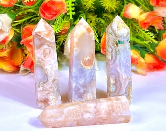 May include: Four agate crystal towers and one rectangular crystal. The crystals have a mix of peach, white, and lavender colours with unique patterns. The crystals are displayed in front of a backdrop of flowers.