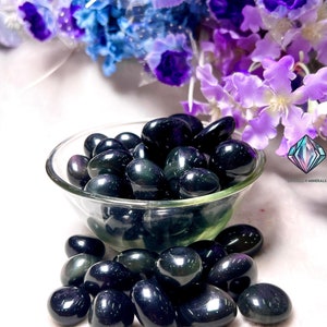May include: A bowl filled with black obsidian tumbled stones. The stones have a dark, iridescent sheen and are arranged in a random pattern. The bowl is sitting on a white surface.