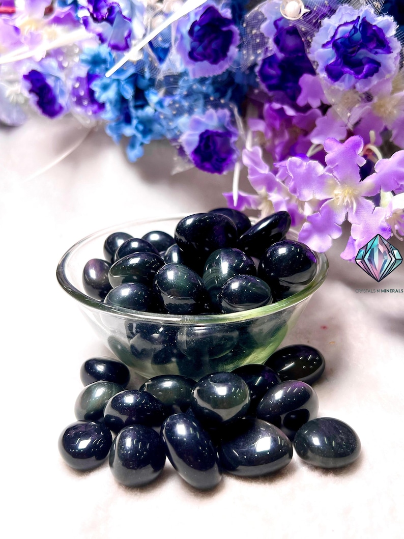 May include: A bowl filled with black obsidian tumbled stones. The stones have a dark, iridescent sheen and are arranged in a random pattern. The bowl is sitting on a white surface.