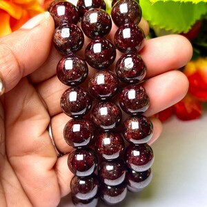 May include: Three red garnet gemstone bracelets with round beads. The bracelets are stacked on top of each other and are being held in a hand.