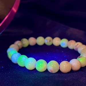 May include: A beaded bracelet with round, light-colored beads. Some beads glow with a bright green and blue hue, while others have a pale pinkish tone. The bracelet is set against a dark background, with a pink object partially visible in the upper left corner.