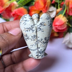 May include: A white and black speckled stone angel figurine carved in the shape of a heart. The angel is standing on a white surface.