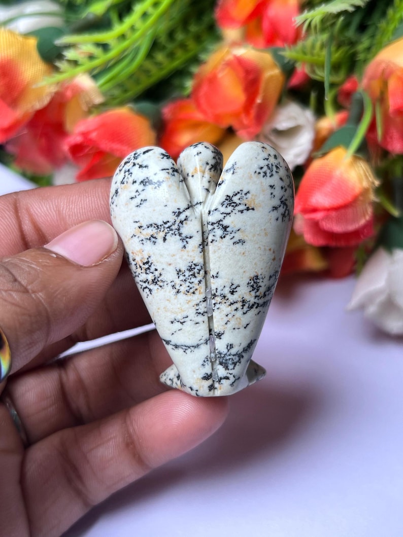 May include: A white and black speckled stone angel figurine carved in the shape of a heart. The angel is standing on a white surface.