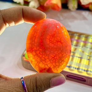 Peut inclure: Une pierre d'agate rouge lisse de forme ovale avec un motif blanc et orange. La pierre est translucide et a un aspect naturel et organique.