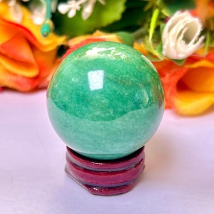 Puede incluir: Una esfera de aventurina verde sobre un soporte de madera roja. La esfera es lisa y pulida, con un ligero brillo.