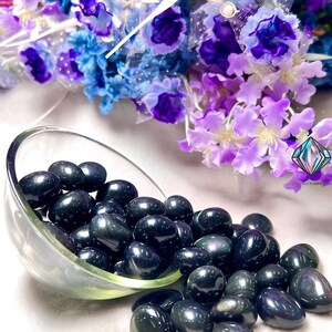 May include: A bowl filled with black obsidian tumbled stones, with a few spilling out onto a white surface. The stones have a dark, iridescent sheen.