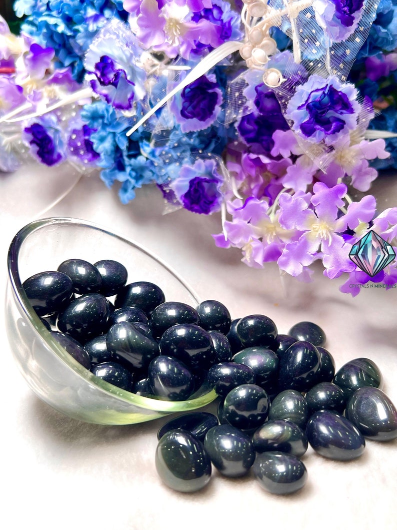 May include: A bowl filled with black obsidian tumbled stones, with a few spilling out onto a white surface. The stones have a dark, iridescent sheen.