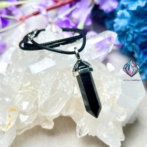 Puede incluir: P&eacute;ndulo de punta de obsidiana negra en un collar de cord&oacute;n negro. El colgante est&aacute; engastado en un bisel de tono plateado y cuelga sobre un fondo de cristales blancos.
