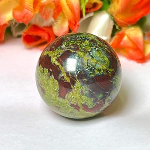 May include: A green, red, and brown polished sphere made of a natural stone. The sphere is smooth and has a glossy finish.