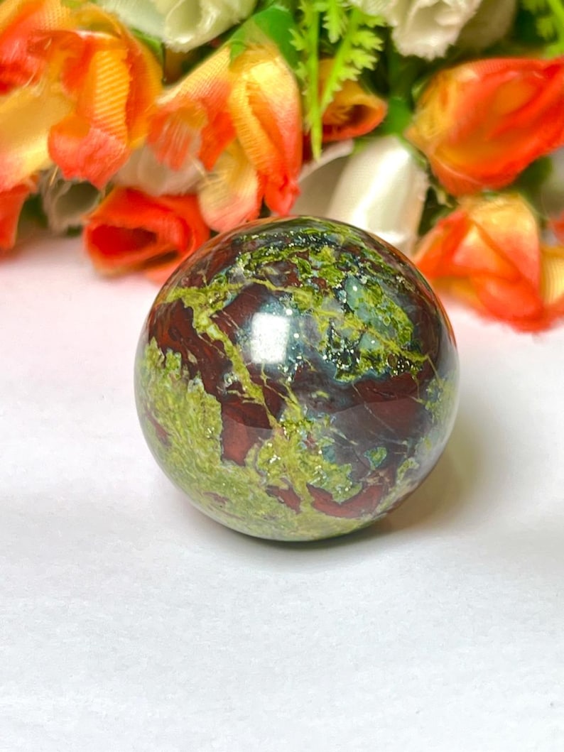 May include: A green, red, and brown polished sphere made of a natural stone. The sphere is smooth and has a glossy finish.