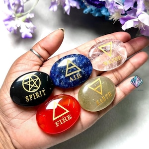 May include: Five smooth, oval-shaped stones with gold symbols and text. The stones are in the colors black, blue, clear, green, and red. The text on the stones reads "Spirit", "Air", "Water", "Earth", and "Fire".