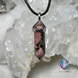 May include: A pink and black rhodochrosite crystal pendant on a black cord necklace. The pendant is set in a silver bezel and has a pointed shape.