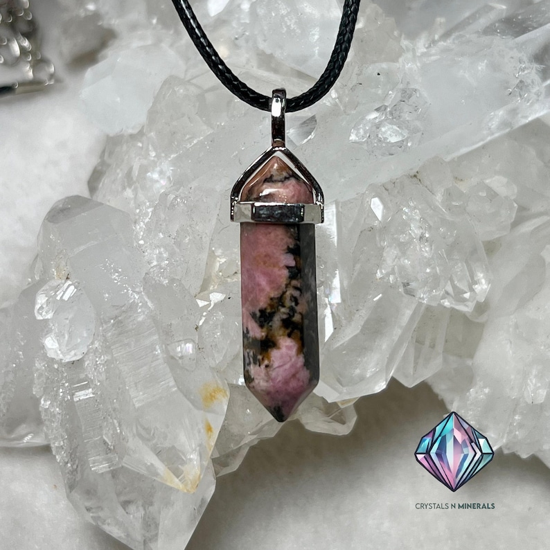 May include: A pink and black rhodochrosite crystal pendant on a black cord necklace. The pendant is set in a silver bezel and has a pointed shape.