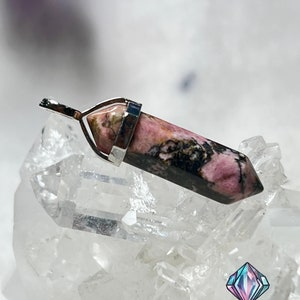 May include: A pink and black rhodochrosite crystal pendant with a silver bail. The pendant is sitting on a bed of clear quartz crystals.