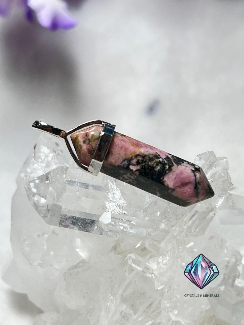 May include: A pink and black rhodochrosite crystal pendant with a silver bail. The pendant is sitting on a bed of clear quartz crystals.