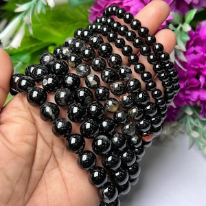 May include: A stack of black gemstone bracelets with silver flecks and red inclusions. The bracelets are made of round beads and are stacked on a hand.