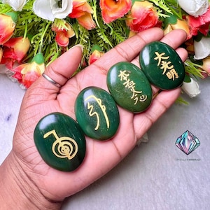 May include: Five green oval-shaped stones with gold symbols inscribed on them. The stones are held in a hand. The symbols are in Chinese characters.