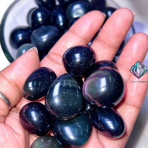 May include: A collection of tumbled obsidian stones, also known as black obsidian, with a dark, glassy appearance. The stones are smooth and polished, with a slight iridescent sheen.
