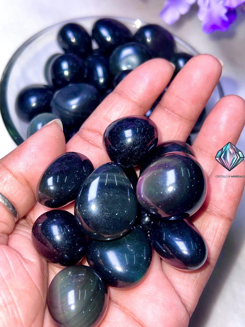 May include: A collection of tumbled obsidian stones, also known as black obsidian, with a dark, glassy appearance. The stones are smooth and polished, with a slight iridescent sheen.
