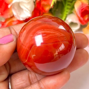 May include: A smooth, polished red carnelian sphere with swirling patterns. The sphere is held in a hand.