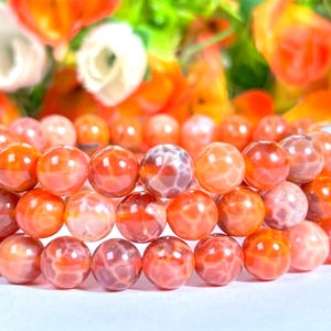Peut inclure: Trois bracelets superposés composés de perles rondes en pierres précieuses orange-rouge. Les perles présentent un aspect marbré avec des variations de couleur. L'arrière-plan est flou, avec des fleurs orange et blanches.