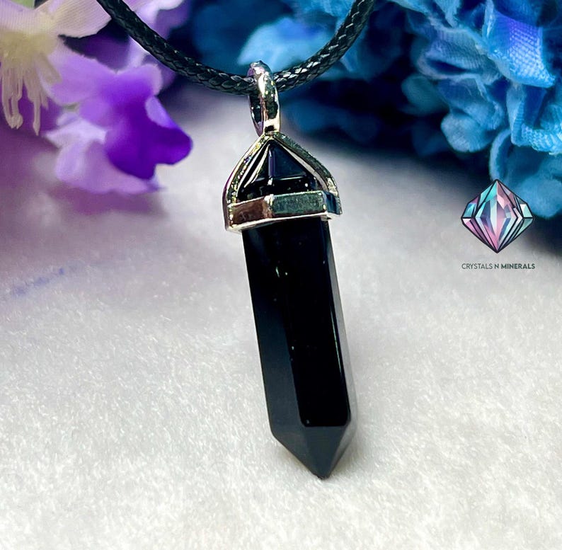 Puede incluir: Colgante de punta de cristal de obsidiana negra en un collar de cord&oacute;n negro. El colgante est&aacute; engastado en un bisel plateado con un peque&ntilde;o cristal en la parte superior.