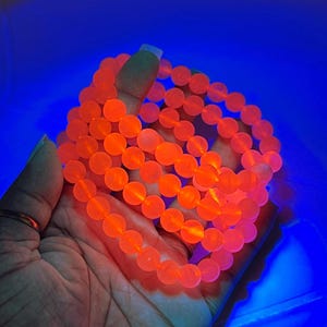May include: A collection of orange beaded bracelets. The bracelets are made of round beads and are illuminated with a bright orange glow against a blue background. The bracelets are held in a hand.